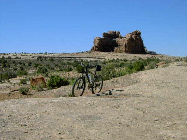 A mountain bike leaning against a barren rock surface, with a large rock formation in the background and sparse vegetation in the surrounding landscape under a clear blue sky. Monitor &amp; Merrimac/Courthouse Pasture mountain bike trail.