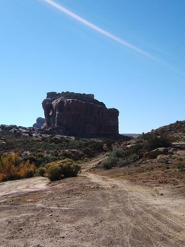 A prominent rock formation rises against a clear blue sky, surrounded by a desert landscape featuring shrubs and dirt pathways. The scene captures the rugged beauty of nature in a remote area. Monitor &amp; Merrimac/Courthouse Pasture mountain bike trail.