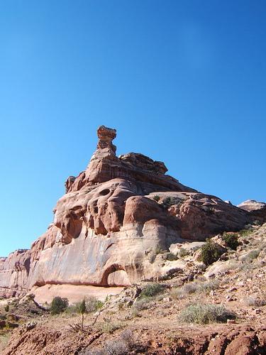 A rocky formation with a prominent, balanced rock at its peak, surrounded by a clear blue sky and vegetation at the base. The landscape features various shades of red and brown in the rock surface. Monitor &amp; Merrimac/Courthouse Pasture mountain bike trail.