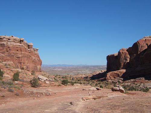 A wide landscape featuring towering red rock formations on either side, with a clear blue sky above and a dusty, barren ground in the foreground. The scene captures the expansive desert terrain with distant mountains visible on the horizon. Monitor &amp; Merrimac/Courthouse Pasture mountain bike trail.