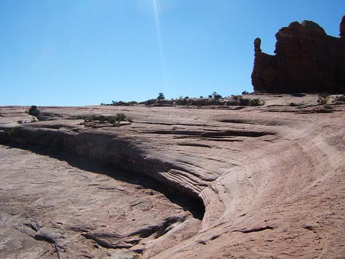 A rugged landscape featuring smooth, sloped rock formations under a clear blue sky. The foreground shows a wide expanse of weathered rock with subtle curves and lines, while the background features a towering rock formation. Sparse vegetation is visible throughout the scene. Monitor &amp; Merrimac/Courthouse Pasture mountain bike trail.