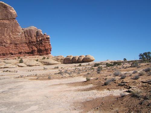 Desert landscape featuring large sandstone rock formations under a clear blue sky, with sparse vegetation and rocky terrain visible in the foreground. Monitor &amp; Merrimac/Courthouse Pasture mountain bike trail.