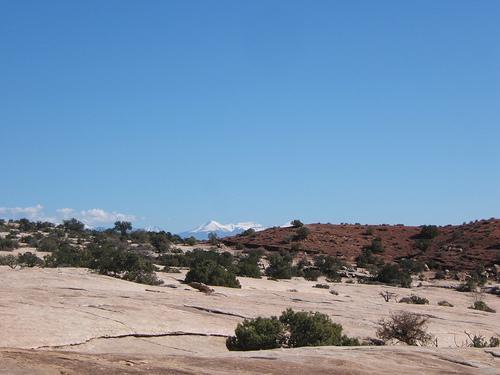 A rocky desert landscape under a clear blue sky, featuring rugged terrain with sparse vegetation and distant snow-capped mountains on the horizon. Monitor &amp; Merrimac/Courthouse Pasture mountain bike trail.