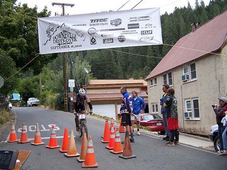 A mountain biker navigates a course marked by traffic cones under a banner announcing the "Bonnieville Destroyer" event, with spectators watching nearby. The background features trees and houses, indicating a rural setting. Downieville Downhill mountain bike trail.