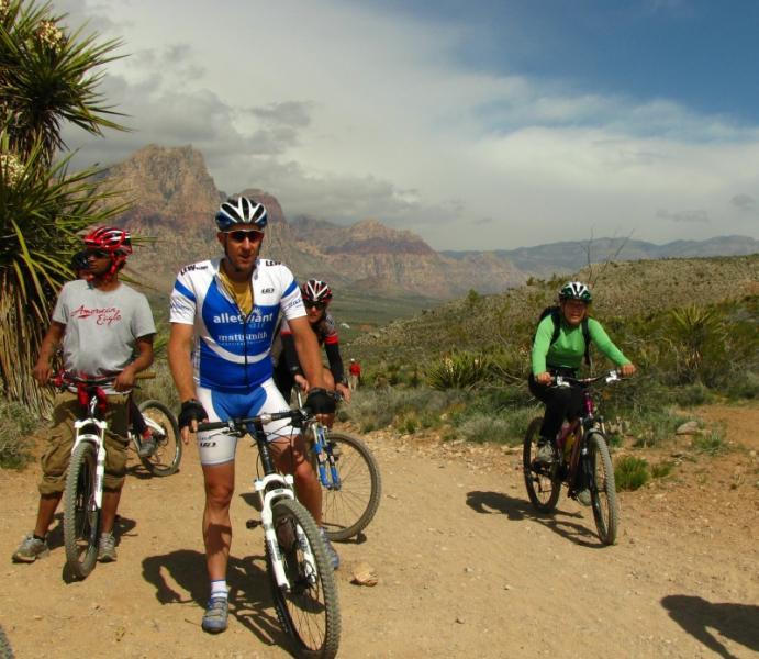 A group of four mountain bikers on a dirt trail in a scenic landscape, featuring tall greenery and rocky mountains in the background. One rider is in a blue and white jersey, while others wear casual and athletic clothing. The sky is partly cloudy, indicating a bright day for outdoor activity. Blue Diamond mountain bike trail.