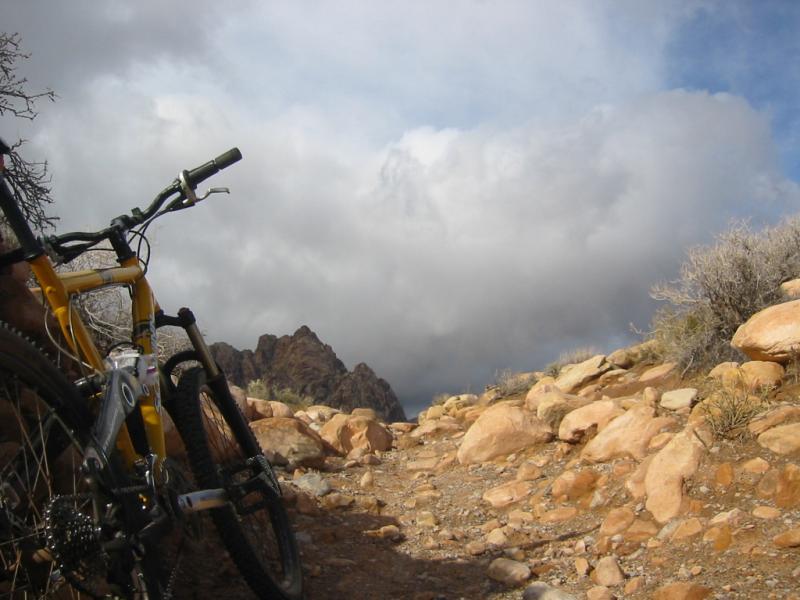 A mountain bike parked on a rocky trail, with a dramatic sky filled with clouds in the background. Surrounding terrain features large stones and sparse vegetation, leading toward rugged mountain peaks. Blue Diamond mountain bike trail.