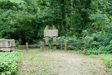 A wooden signpost labeled "Jean-Pierre Chouteau Nature Trail" is seen at the entrance of a natural area, surrounded by lush green foliage. The trail is marked with a fence that restricts motorized vehicle access. The pathway is visible, leading into the wooded area. Jean-pierre Chouteau Trail mountain bike trail.