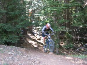 A mountain biker navigating a narrow trail through a wooded area filled with trees and sunlight filtering through the branches. Northstar Bike Park mountain bike trail.