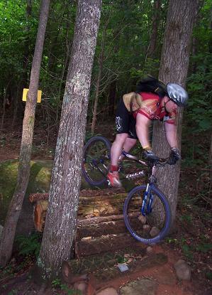 A mountain biker navigating a rocky trail, performing a jump over a wooden feature while surrounded by trees and lush green foliage. The rider is wearing a helmet and protective gear, demonstrating skill and agility on the bike. Fisher Farm Park mountain bike trail.