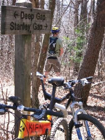 Mountain bikers resting at a trail junction marked by a wooden sign pointing to "Deep Gap" and "Stanley Gap," with a few mountain bikes in the foreground and a rider standing in the background among trees. Stanley Gap mountain bike trail.