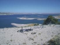 A bicycle rests on a rocky shore overlooking a vast, blue lake with small islands in the distance under a clear blue sky. South Shore Lake Pueblo mountain bike trail.