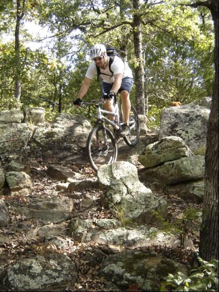 A mountain biker navigating a rocky trail surrounded by trees. The cyclist is wearing a helmet and a backpack, focused on maintaining balance while riding over the uneven terrain. Sunlight filters through the leaves, creating a natural, outdoor setting. Lake Murray State Park mountain bike trail.