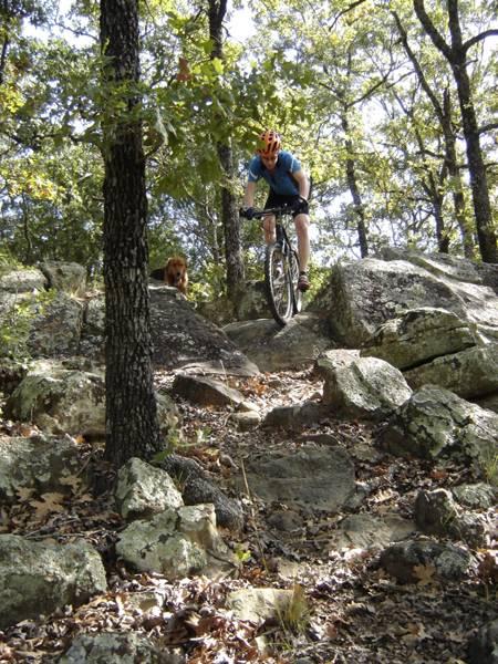 A mountain biker navigating over rocky terrain with trees in the background, accompanied by a dog watching nearby. Lake Murray State Park mountain bike trail.