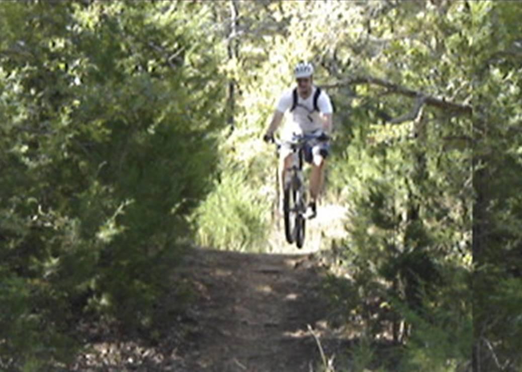 A person in a white shirt and helmet is riding a mountain bike on a narrow dirt trail surrounded by green foliage, showing a moment of airborne action as they jump. Lake Murray State Park mountain bike trail.