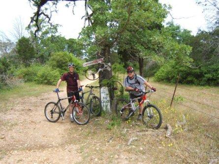 Two mountain bikers standing beside their bikes at a trail intersection. The scene includes a tree with greenery around, and a sign indicating trail directions. The bikers are wearing helmets and casual cycling attire. Rocky Hill Ranch mountain bike trail.