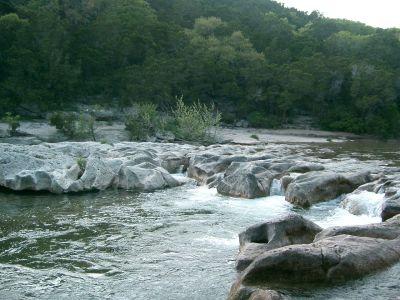 A serene river flowing over rocky formations, surrounded by lush greenery and trees in the background. The water cascades gently over the stones, creating a tranquil natural scene. Barton Creek Greenbelt mountain bike trail.