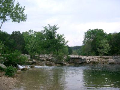 A tranquil scene of a river surrounded by lush greenery and rocky outcrops, with a cloudy sky overhead. The calm water reflects the trees and rocks, creating a serene natural setting. Barton Creek Greenbelt mountain bike trail.