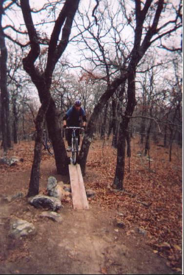 A mountain biker in mid-air, jumping off a wooden ramp positioned between two trees in a forested area with fallen leaves on the ground. Lake Murray State Park mountain bike trail.