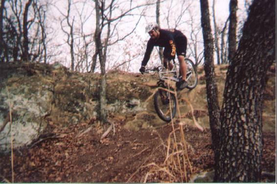 A person riding a mountain bike jumps off a rock ledge in a wooded area with bare trees in the background. The ground is covered with fallen leaves, and the biker is wearing gear suitable for off-road cycling. Lake Murray State Park mountain bike trail.