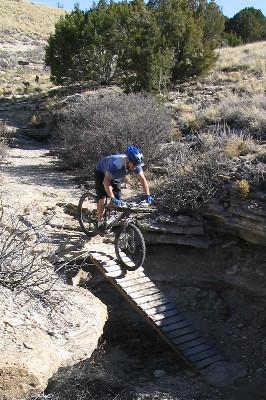A mountain biker in a blue helmet navigates a narrow wooden bridge over rocky terrain, surrounded by sparse vegetation and hills. The scene captures a moment of action and skill in an outdoor biking environment. South Shore Lake Pueblo mountain bike trail.