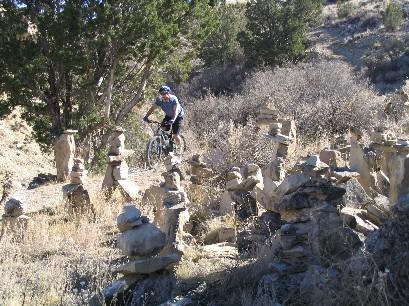 A mountain biker rides through a rocky landscape filled with stacked stone cairns, surrounded by low brush and trees on a sunny day. South Shore Lake Pueblo mountain bike trail.