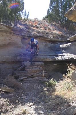 A mountain biker performing a jump over a rocky terrain, with rugged cliffs and shrubbery in the background. The scene captures the thrill of outdoor biking in a natural landscape. South Shore Lake Pueblo mountain bike trail.