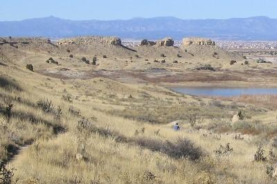 A serene landscape featuring rolling hills covered in dry grass and scattered shrubs, with a small lake in the foreground. The distant mountains are visible under a clear blue sky. A lone figure can be seen walking along a trail in the lower part of the image. South Shore Lake Pueblo mountain bike trail.