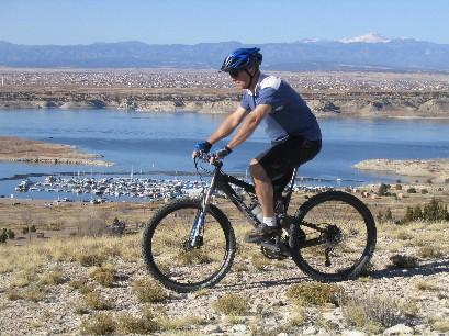 A mountain biker rides a black bike on a rocky hillside, overlooking a lake with boats and a distant mountainous landscape under a clear blue sky. South Shore Lake Pueblo mountain bike trail.