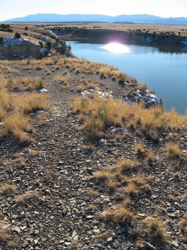 A rocky trail leading toward a calm, reflective body of water, surrounded by sparse vegetation and rolling hills in the background, under a clear blue sky. South Shore Lake Pueblo mountain bike trail.