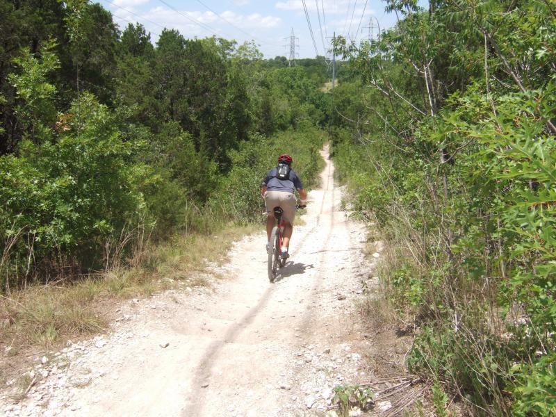 A person riding a mountain bike along a dirt trail, surrounded by lush greenery and trees, under a bright blue sky. The trail winds through a natural landscape, with power lines visible in the distance. Walnut Creek Trails mountain bike trail.