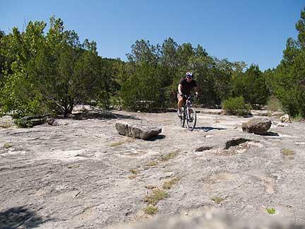 A mountain biker navigating a rocky trail surrounded by sparse trees and clear blue skies. Barton Creek Greenbelt mountain bike trail.