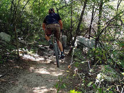 A person riding a mountain bike on a narrow dirt trail surrounded by trees and rocks, with sunlight filtering through the foliage. Barton Creek Greenbelt mountain bike trail.