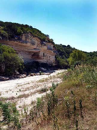 A scenic view of a dry riverbed bordered by steep cliffs and lush vegetation, under a clear blue sky. The area showcases rocky terrain and sparse grasses, indicating a natural landscape. Barton Creek Greenbelt mountain bike trail.