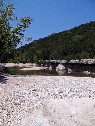 A serene outdoor scene featuring a shallow stream bordered by smooth stones and pebbles. Lush greenery and trees line the banks, with a rocky formation visible in the background under a clear blue sky. Barton Creek Greenbelt mountain bike trail.