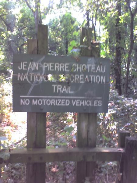A wooden sign for the Jean Pierre Choteau National Recreation Trail, indicating that no motorized vehicles are allowed. The sign is surrounded by lush greenery in a wooded area. Jean-pierre Chouteau Trail mountain bike trail.