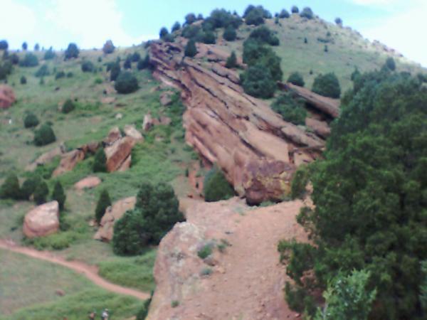 A rocky hillside with a prominent rock formation, surrounded by green vegetation and trees. A winding dirt path leads through the landscape. The sky above features a few clouds. Matthews Winters mountain bike trail.