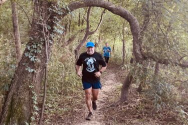 Two people are jogging on a dirt trail surrounded by trees and greenery. One runner, wearing a black shirt and blue shorts, is in the foreground, while a second runner, dressed in a blue shirt, is in the background. A distinctive curved tree branches above the trail, adding to the natural scenery. Jean-pierre Chouteau Trail mountain bike trail.