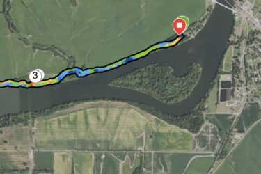 Aerial map showing a winding river bordered by green fields and trees, with marked waypoints along the water's edge. Key points are indicated with circles and labels, while different colors represent various features of the terrain. A prominent red pin marks a specific location near the river. Jean-pierre Chouteau Trail mountain bike trail.