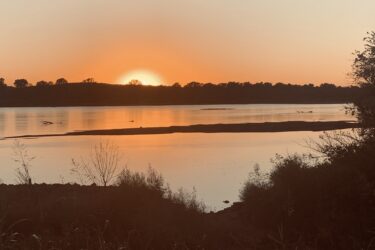 A serene sunset over a calm river, with the sun dipping below the horizon. Silhouettes of trees and shoreline vegetation frame the scene, reflecting soft hues of orange and gold on the water's surface. Jean-pierre Chouteau Trail mountain bike trail.