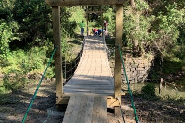 A wooden suspension bridge leads across a small creek, surrounded by lush green vegetation. People can be seen on the far side, engaged in an activity, while the bridge is secured with ropes on either side. The scene features natural elements and a peaceful outdoor setting. Jean-pierre Chouteau Trail mountain bike trail.