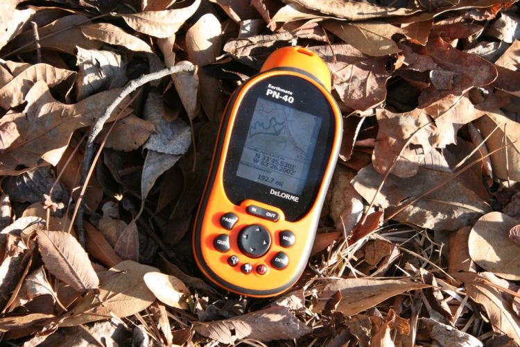An orange GPS device, the Earthmate PN-40, resting on a bed of brown dried leaves, displaying a mapping interface with coordinates and distance information.