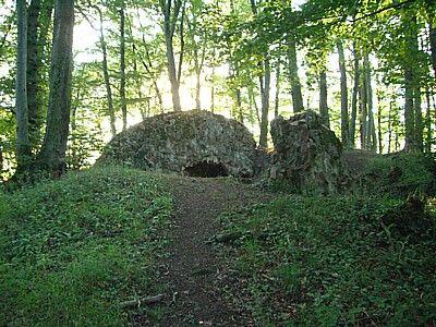 A stone structure resembling a small cave, surrounded by greenery and trees, with sunlight filtering through the leaves in the background. A dirt path leads up to the entrance of the structure. Hirschberg C5 mountain bike trail.