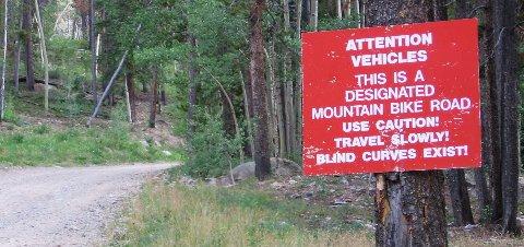 A bright red sign near a dirt road in a wooded area, warning vehicles that it is a designated mountain bike road. The sign instructs to use caution, travel slowly, and notes the presence of blind curves. Keystone Gulch mountain bike trail.
