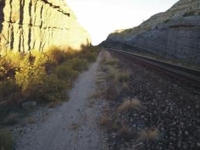 A view of a narrow path alongside railway tracks, flanked by tall rocky cliffs. The sunlight illuminates the scene, highlighting the textures of the rocks and the scattered vegetation along the path. PMI / Nature Trails mountain bike trail.