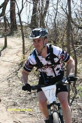 A mountain biker wearing a helmet and a colorful cycling jersey navigates a dirt trail surrounded by trees. The cyclist is focused and mid-action, with a race number displayed on their jersey. Sunlight filters through the trees, creating a natural outdoor setting. Lawrence Riverfront Trails mountain bike trail.