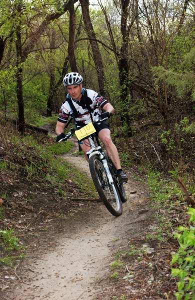 A cyclist with a race number is riding a mountain bike on a dirt trail surrounded by greenery and trees. The rider is leaning into a turn, showcasing an action-packed moment during a mountain biking event. Lawrence Riverfront Trails mountain bike trail.