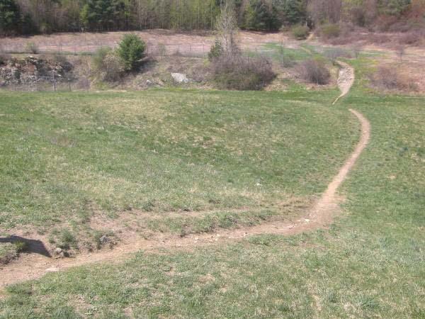 A winding dirt path through a grassy field, surrounded by trees and a gentle hillside. The scene captures the natural landscape with patches of greenery, leading into the distance where the terrain changes. Callahan State Park mountain bike trail.