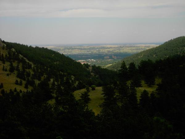 A panoramic view of a valley surrounded by rolling hills and mountains, with lush green trees in the foreground and a distant landscape stretching out under a partly cloudy sky. Betasso Preserve mountain bike trail.