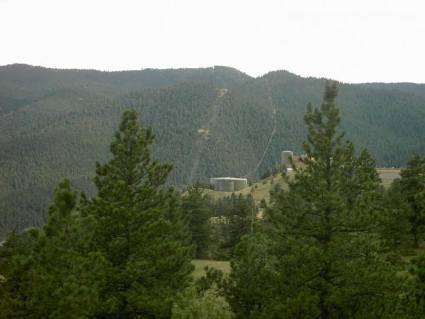 A panoramic view of a mountainous landscape featuring green pine trees in the foreground, with rolling hills and a distant structure visible in the middle ground. The sky is overcast, creating a serene, natural setting. Betasso Preserve mountain bike trail.