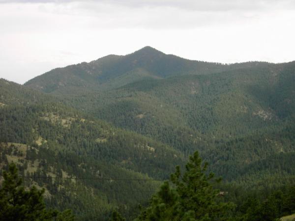 A scenic view of a mountainous landscape featuring a prominent peak surrounded by dense forests and rolling hills under a cloudy sky. Betasso Preserve mountain bike trail.